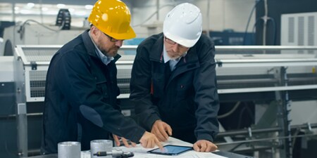 Two people wearing hard hats look at a tablet risting on a table.
