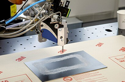 A robot arm applies adhesive to a metal plate in a rectangular pattern