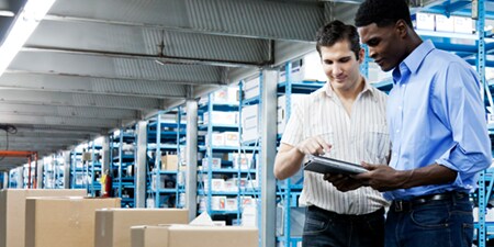 Two people standing in front of shelves in a warehouse look at a document.
