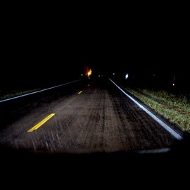 Vehicle headlights illuminate a rainy road in the dark, with visible pavement markings and an orange road sign ahead