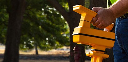 A man walks near a forested area carrying a 3M tool used for locating, marking and mapping underground assets.
