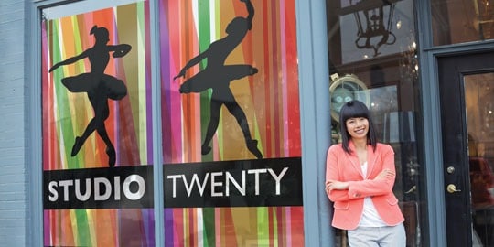 A woman leans against a storefront with large, bold window wraps displaying dancers and the words &ldquo;studio twenty.&rdquo;